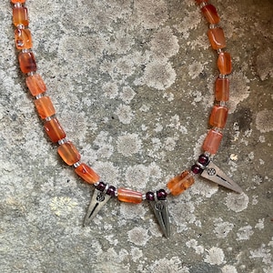 May include: A necklace featuring orange-red, rectangular beads strung together with smaller clear beads. The necklace has three silver-colored, triangular pendants with decorative details, and small, dark red beads are interspersed near the pendants.