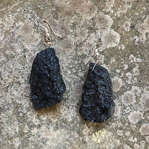 May include: A pair of black, textured earrings with silver-colored hooks. The earrings have a rough, organic shape, and are displayed against a mottled gray and tan background.