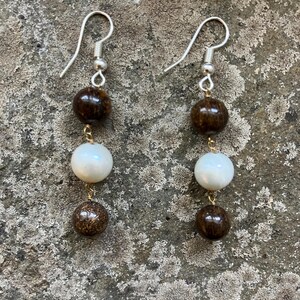 May include: A pair of dangle earrings featuring a combination of dark brown and white beads. The earrings have a silver hook and are connected by gold-colored wire. The beads have a glossy finish and are arranged in a vertical line.