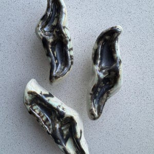 May include: Three abstract ceramic sculptures with a glossy finish. The pieces have a cream base with dark brown and black accents. The forms are organic and curved, resembling stylized leaves or pods.