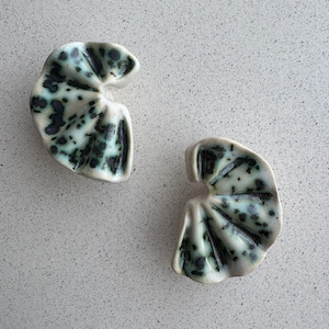 May include: Two ceramic leaf-shaped dishes. The dishes are white with a speckled pattern of teal and dark blue. The leaves have a folded, textured design. The dishes are placed on a speckled, light grey surface.