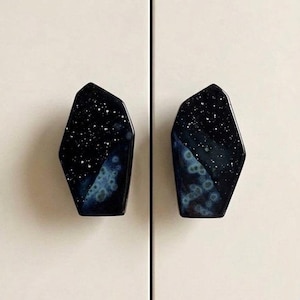 May include: Two asymmetrical cabinet knobs with a glossy black finish featuring a speckled white star pattern and a blue nebula-like glaze effect. The knobs have a faceted, geometric shape and are mounted on a smooth, light-colored surface.