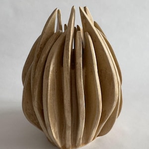 May include: A beige, handcrafted decorative vase with a unique, organic design. The vase features a series of vertical, petal-like elements that curve inwards, creating a textured surface. The overall shape is reminiscent of a blooming flower or a stylized bud.