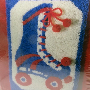 Puede incluir: Un kit de alfombra de gancho con un diseño de patín de ruedas azul y rojo con ruedas blancas. El kit incluye pompones y el texto "latch hook with a new twist!"