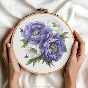 Puede incluir: Un bastidor de bordado de punto de cruz con un diseño floral de peonías moradas y blancas con hojas verdes. El bordado está sobre un fondo de tela blanca. El bastidor de madera está sostenido por dos manos.