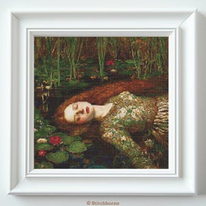 May include: A framed cross-stitch of a woman in a pond, based on the painting Ophelia. The woman has long red hair and wears a green dress. The scene includes water lilies and tall reeds, all within a white frame.