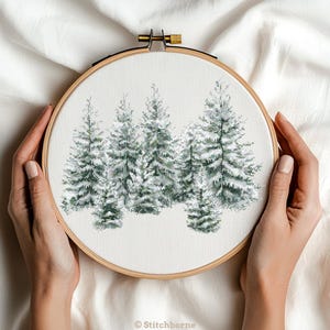 May include: A cross-stitch embroidery hoop featuring a winter forest scene. The design showcases snow-covered evergreen trees in shades of green and white. The embroidery is set in a wooden hoop, held by hands against a white fabric backdrop. The text "Stitchborne" is at the bottom.