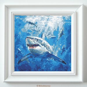 May include: A framed cross-stitch artwork depicts a great white shark swimming in blue water, surrounded by smaller fish. The shark is detailed with open jaws and visible teeth. The artwork is framed in a white square frame.