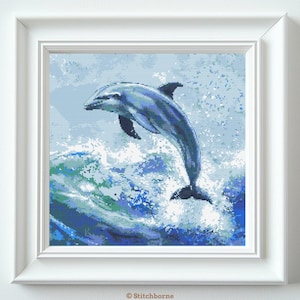 May include: A framed cross-stitch artwork depicts a leaping dolphin in shades of blue and white. The dolphin is mid-air, above a crashing wave, with a light blue background. The artwork is in a white frame.