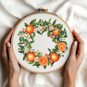 May include: Cross-stitch embroidery of a wreath featuring oranges, white blossoms, and green leaves. The design is set in a wooden embroidery hoop. The oranges are a vibrant orange colour, and the leaves are a deep green. The embroidery is on a white fabric.
