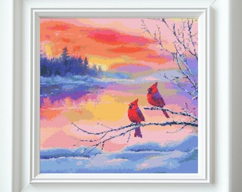 May include: Framed cross-stitch art featuring two red cardinals perched on a snow-covered branch. The background depicts a colorful sunset over a lake and forest, with hues of orange, pink, and blue. The artwork is in a white frame.