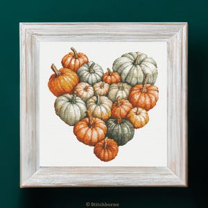 May include: A framed cross-stitch artwork featuring a heart shape composed of various pumpkins. The pumpkins are in shades of orange, white, and green, with textured surfaces and green stems. The frame is a distressed white color.