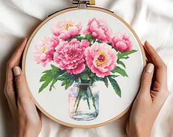 May include: Cross-stitch embroidery of pink peonies in a glass vase, set in a wooden hoop. The flowers have yellow centers and green leaves. The embroidery is held by hands against a white fabric background. The text "Stitchborne" is at the bottom.