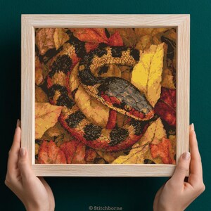 Könnte beinhalten: Kreuzstich-Kunstwerk einer Schlange, die sich zwischen Herbstblättern windet, in einem Holzrahmen. Die Schlange hat ein schwarz-rotes Muster mit einem gelb-schwarzen Kopf. Die Blätter sind in Gelb-, Orange- und Rottönen gehalten.
