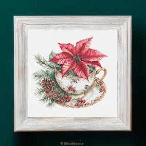 Può includere: Opera d'arte a punto croce in una cornice di legno bianca. L'immagine presenta una tazza da tè piena di una stella di Natale rossa, aghi di pino e pigne. La tazza e il piattino hanno un motivo floreale. L'opera è su uno sfondo verde scuro.