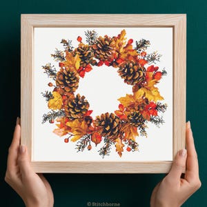 May include: A framed cross-stitch wreath featuring autumn elements. The wreath is composed of pinecones, fall leaves in shades of orange and yellow, and red berries. The frame is light-colored wood, and the background is white.
