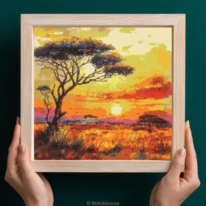 Op de afbeelding: Een ingelijst kruissteeklandschap dat een Afrikaanse savanne bij zonsondergang uitbeeldt. Het kunstwerk toont een grote boom tegen een oranje en gele lucht. De voorgrond toont hoog, goudkleurig gras.