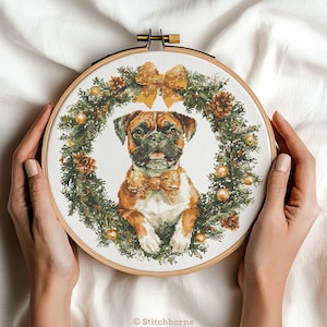May include: A cross-stitch embroidery hoop featuring a Boxer dog wearing a bow tie, surrounded by a Christmas wreath with pinecones, ornaments, and a gold bow. The dog is brown, white, and black. The hoop is held by hands against a white background. The text "© Stitchborne" is at the bottom.