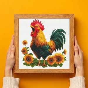 May include: A framed cross-stitch of a rooster with a red comb and open beak, surrounded by sunflowers. The rooster has yellow, orange, and green plumage. The artwork is in a wooden frame, held by hands against a yellow background.