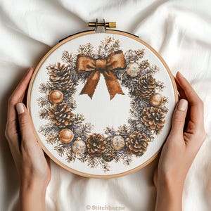 May include: A cross-stitch Christmas wreath design in a wooden embroidery hoop. The wreath features pinecones, ornaments, and a brown bow. The design is stitched on white fabric. The text "© Stitchborne" is at the bottom.