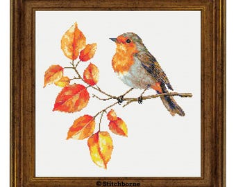 May include: Cross-stitch artwork of a robin perched on a branch with orange and yellow leaves. The bird has a red breast and brown and gray feathers. The artwork is framed in a brown wooden frame. The text "Stitchborne" is in the bottom corner.