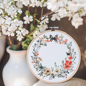 Puede incluir: Un aro de bordado blanco con un diseño de corona floral que incluye mariposas, flores rojas y hojas verdes. La corona está rodeada de un fondo blanco.