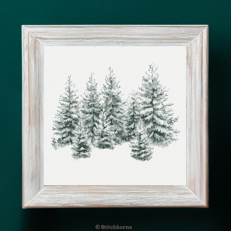 May include: A framed cross-stitch artwork featuring a winter forest scene. The image shows several snow-covered evergreen trees in shades of green and white. The artwork is set in a rustic, light-colored wooden frame, perfect for home decor.