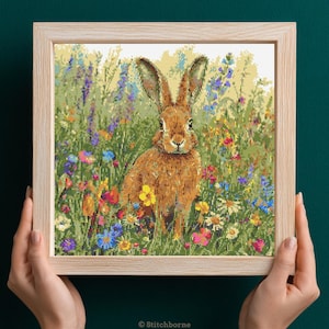 Puede incluir: Un bordado a punto de cruz enmarcado de una liebre marrón en un campo de flores silvestres coloridas. La liebre tiene orejas largas y está rodeada de flores azules, amarillas, rosas y rojas. El marco es de madera clara.