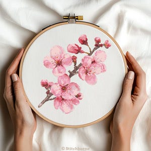 May include: A cross-stitch embroidery of pink cherry blossoms on a branch, displayed in a wooden hoop. The floral design features detailed petals and buds, with a brown stem. The hoop is held by hands, and the background is a white fabric.