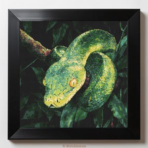 May include: A framed cross-stitch artwork depicting a green snake coiled on a branch amidst green leaves. The snake has yellow eyes and a detailed texture. The artwork is presented in a black square frame, creating a striking contrast.