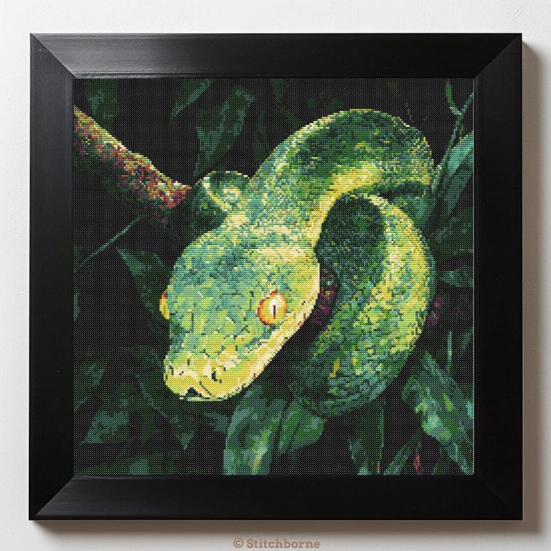 May include: A framed cross-stitch artwork depicting a green snake coiled on a branch amidst green leaves. The snake has yellow eyes and a detailed texture. The artwork is presented in a black square frame, creating a striking contrast.