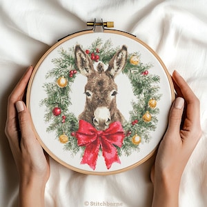 May include: A cross-stitch embroidery hoop featuring a donkey inside a Christmas wreath. The wreath is made of green pine needles, red berries, and gold and red ornaments. A large red bow is at the bottom. The embroidery is held by a wooden hoop.