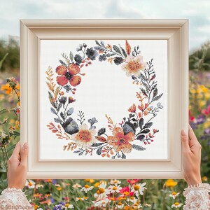 Puede incluir: Un patrón de punto de cruz de una corona floral con fondo blanco. La corona está hecha de flores y hojas de color naranja, rosa y azul.