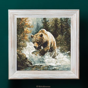 Puede incluir: Un bordado enmarcado que representa un oso pardo en un río, persiguiendo a un pez. El oso es marrón con reflejos blancos, y el agua es blanca y azul. El fondo presenta árboles en tonos verdes y dorados. El marco es blanco.