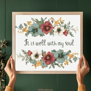 May include: Cross stitch pattern featuring a floral wreath with red, white, and blue flowers. The text "It is well with my soul" is centered below the wreath.