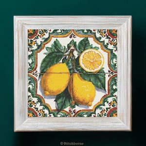 Puede incluir: Obra de arte enmarcada con un diseño de punto de cruz de limones. Los limones amarillos con hojas verdes se encuentran sobre un fondo de azulejos decorativos con patrones verdes, blancos y naranjas. El marco es de madera blanqueada.