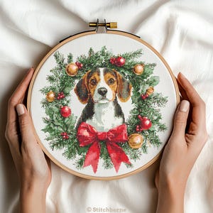 May include: A cross-stitch embroidery hoop featuring a beagle dog surrounded by a Christmas wreath. The wreath includes green pine needles, red berries, and gold and red ornaments. A large red bow is at the bottom. The hoop is held by hands.