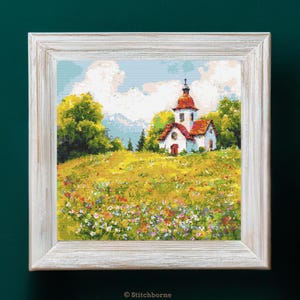 Puede incluir: Un bordado enmarcado representa una iglesia blanca con tejado rojo sobre una colina herbosa salpicada de flores silvestres. El cielo es azul con nubes blancas. El marco es blanco con un acabado envejecido. La imagen está firmada "© Stitchborne".