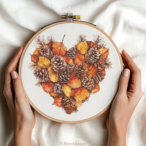 Peut inclure: Broderie au point de croix dans un cercle en bois, représentant un cœur composé de feuilles d'automne et de pommes de pin aux tons orange, jaune et marron. La broderie est tenue par deux mains. Le texte "Stitchborne" est en bas.