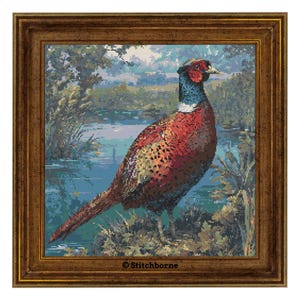 May include: A framed cross-stitch of a pheasant standing near a body of water. The pheasant has red, brown, and yellow feathers, a blue neck, and a white collar. The background shows a landscape with trees and water. The frame is a dark brown color. The text "© Stitchborne" is in the bottom right corner.