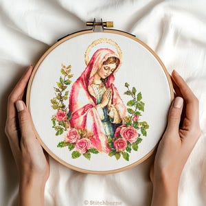 May include: A cross-stitch embroidery of a religious figure with a halo, wearing a pink robe, and surrounded by pink roses and green foliage. The artwork is displayed in a wooden embroidery hoop.