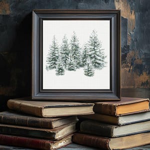 May include: A framed print featuring a watercolor painting of several snow-covered evergreen trees. The artwork is set against a white background and framed in a dark, rustic wooden frame. The frame rests on a stack of antique books.