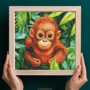 Op de afbeelding: Een kruissteek van een baby orang-oetan met roodbruine vacht, omlijst in licht hout. De orang-oetan is omgeven door groene bladeren. De afbeelding wordt door twee handen vastgehouden.