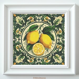 May include: A framed cross-stitch artwork featuring a still life of lemons. Two whole lemons and a sliced lemon are arranged with green leaves on a decorative plate. The background has a green and cream floral pattern. The frame is white.