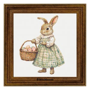 Könnte beinhalten: Kreuzstich eines Hasen in einem grünen karierten Kleid, der einen Korb mit Ostereiern trägt. Der Hase ist braun und weiß, der Korb geflochten. Das Kunstwerk ist in einem braunen Holzrahmen gerahmt.