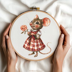 May include: A cross-stitch embroidery hoop with a cute mouse holding a pink flower. The mouse is wearing a red and white checkered dress. The embroidery is on white fabric.