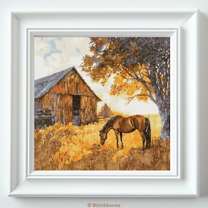 May include: A framed cross-stitch landscape depicts a brown horse grazing in a field near a weathered barn. The scene is bathed in warm, golden light, with a large tree providing shade. The artwork evokes a sense of rural tranquility.