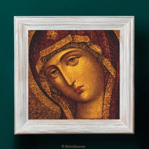 Puede incluir: Icono religioso enmarcado que representa a una mujer con una expresión serena. La obra de arte utiliza tonos cálidos de oro, rojo y marrón. El marco es de color blanco envejecido. La imagen es un diseño de punto de cruz. El texto "© Stitchborne" es visible.
