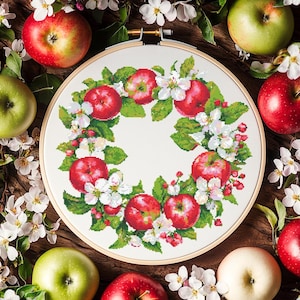 May include: A cross-stitch embroidery hoop featuring a wreath design of red apples, white blossoms, and green leaves. The embroidery is set against a white background, surrounded by fresh red and green apples and white flowers on a wooden surface.