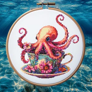 Peut inclure: Broderie au point de croix d'un poulpe dans une tasse à thé. Le poulpe est de couleur corail avec des yeux bleus et des tentacules roses. La tasse et la soucoupe sont décorées de motifs floraux bleus, roses et verts. La broderie est dans un cercle en bois.